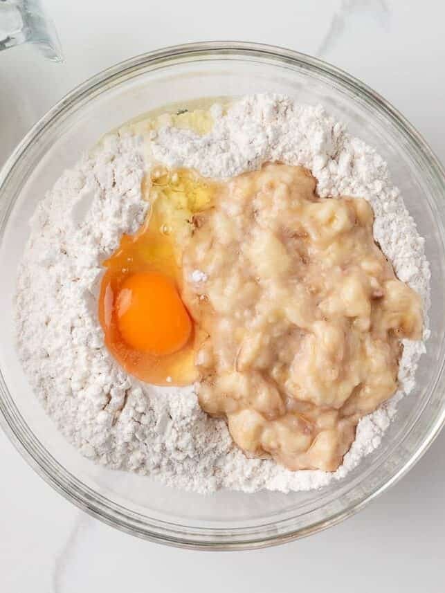 Glass bowl holding flour, mashed banana, and a cracked egg ready to be mixed.