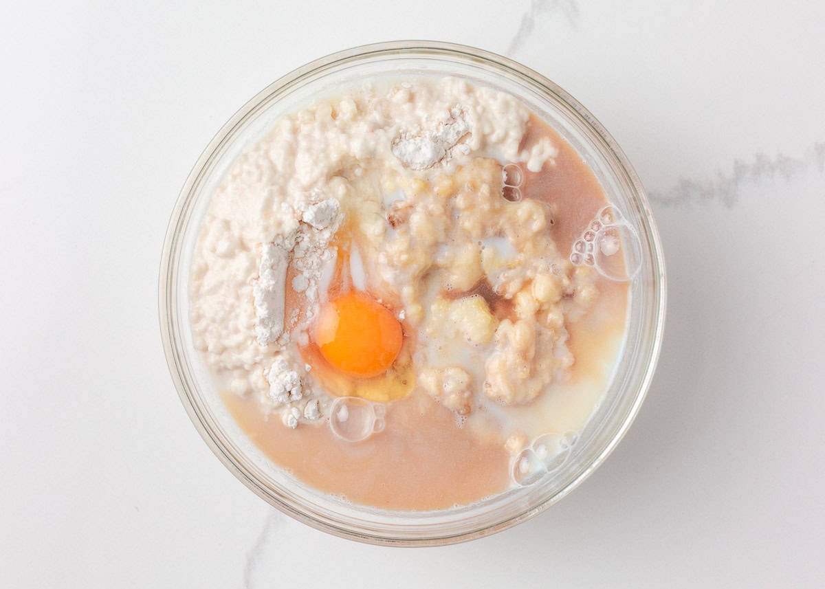 Glass mixing bowl containing pancake muffin batter with mashed banana, flour, milk, a cracked egg, and vanilla before stirring.