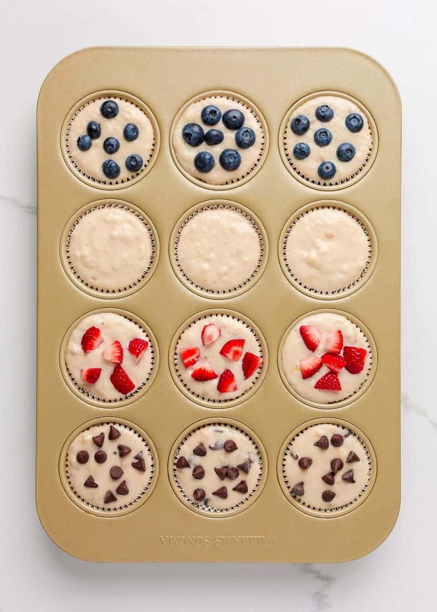 Muffin tin lined with paper liners filled with pancake muffin batter, each topped with blueberries, strawberries, or chocolate chips.