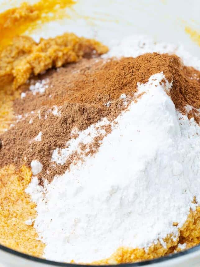 Pumpkin cookie dough in a glass bowl with flour, cinnamon, nutmeg, and baking soda added on top, ready to be mixed.