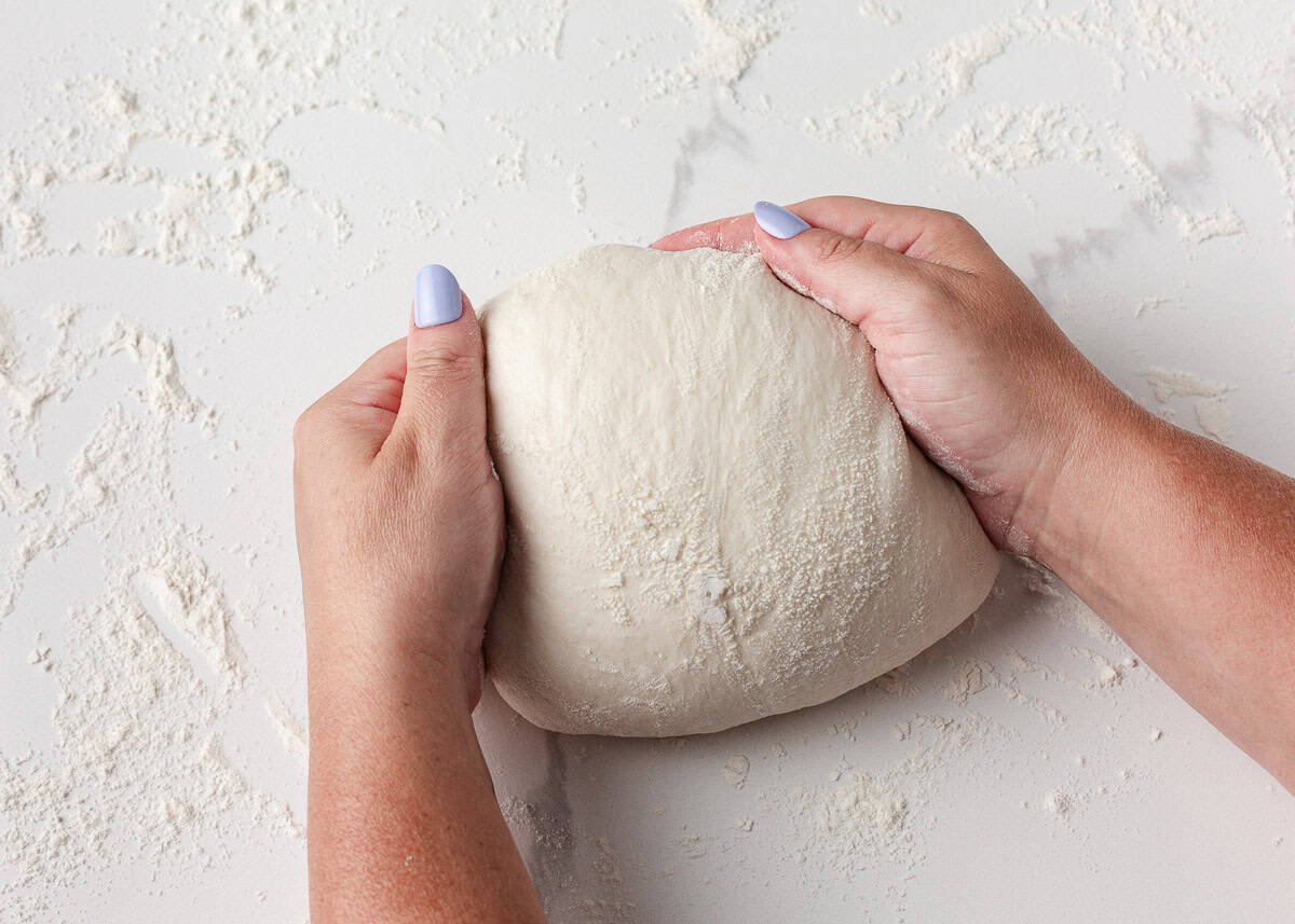Hands gently folding same day sourdough bread dough on a floured surface during shaping into a boule.