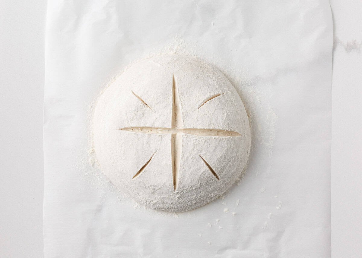 Round loaf of same day sourdough dough scored with a cross and placed on parchment paper, ready to bake.