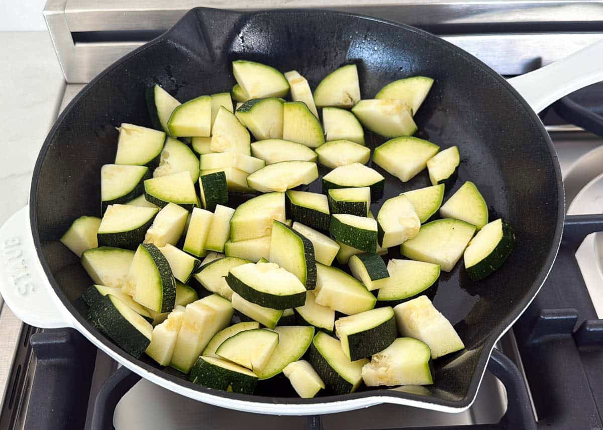 This simple sautéed zucchini is easy to make with a bit of oil, seasoning, and a finish of fresh herbs and Parmesan cheese for the tastiest side dish that the whole family will love! Raw zucchini pieces added to a skillet with olive oil to make sautéed zucchini.