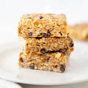 Close-up of soft and chewy no bake oatmeal bars stacked on a white plate with oats and chocolate chips.