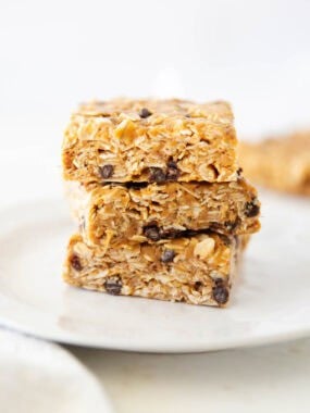Close-up of soft and chewy no bake oatmeal bars stacked on a white plate with oats and chocolate chips.