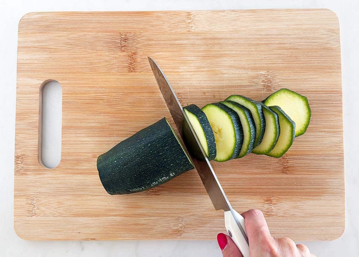 This simple sautéed zucchini is easy to make with a bit of oil, seasoning, and a finish of fresh herbs and Parmesan cheese for the tastiest side dish that the whole family will love! Fresh zucchini being sliced into even rounds on a cutting board for sautéing.