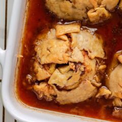 Golden brown apple dumplings nestled in a rich caramel-like sauce, shown in the corner of a white baking dish.