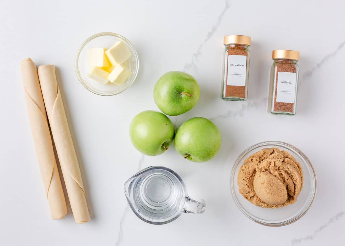 Overhead view of ingredients for apple dumplings including Granny Smith apples, pie crusts, butter, brown sugar, cinnamon, nutmeg, and water on a white surface.