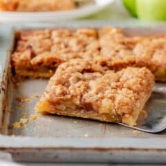 Apple slab pie cut into squares with a spatula lifting out a slice, showing tender apple filling and crumb topping.