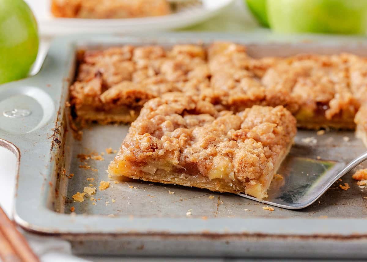 Apple slab pie cut into squares with a spatula lifting out a slice, showing tender apple filling and crumb topping.