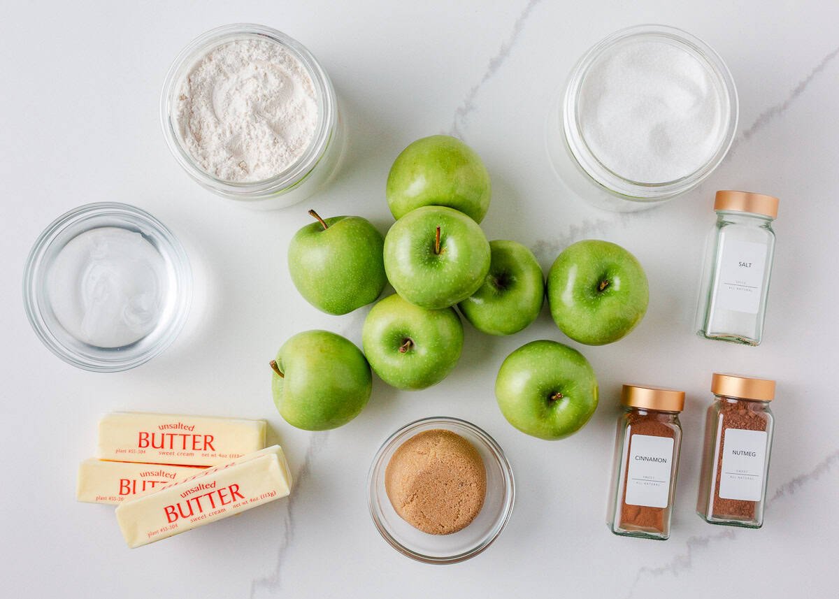 IIngredients for apple slab pie including Granny Smith apples, flour, sugar, butter, brown sugar, cinnamon, nutmeg, salt, and water arranged on a white counter.