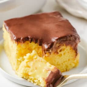 Slice of Boston cream poke cake with chocolate ganache topping and creamy pudding center on a white plate.