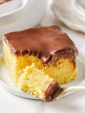 Slice of Boston cream poke cake with chocolate ganache topping and creamy pudding center on a white plate.