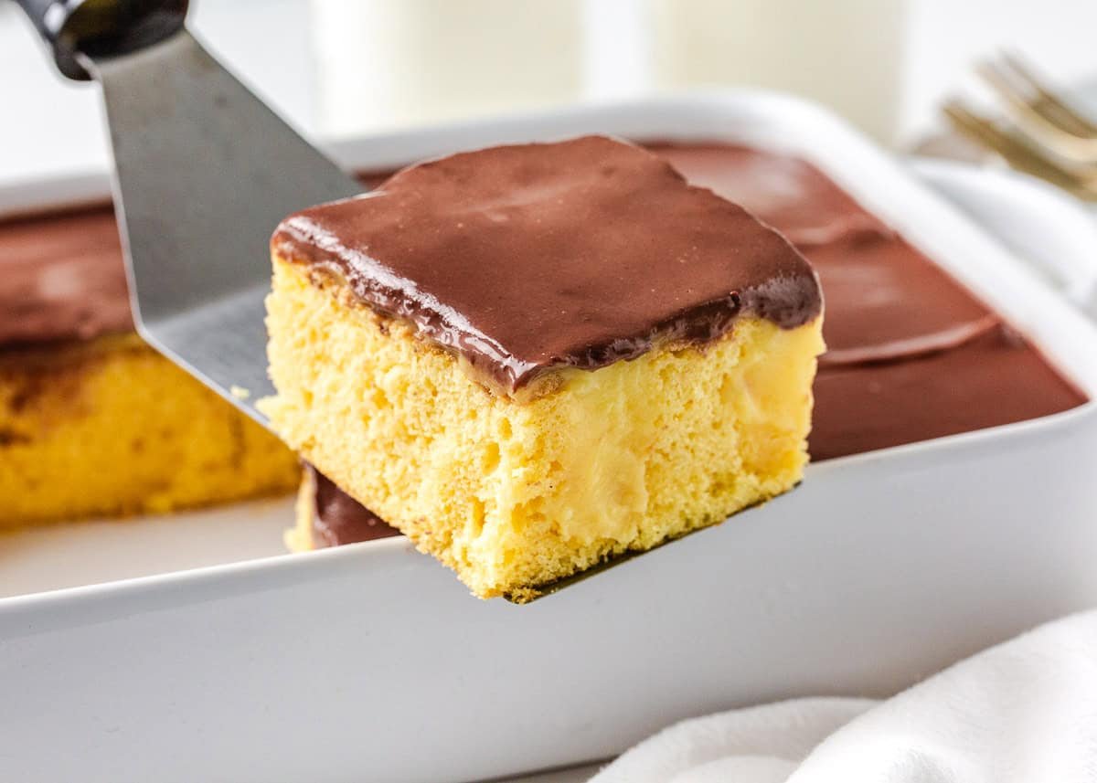 Boston cream poke cake slice being lifted from the pan showing creamy custard filling and chocolate topping.