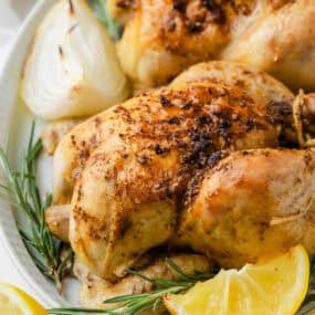 Juicy roasted cornish game hens on a platter with fresh rosemary sprigs, baked onions, and lemon slices.
