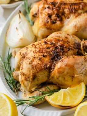 Juicy roasted cornish game hens on a platter with fresh rosemary sprigs, baked onions, and lemon slices.