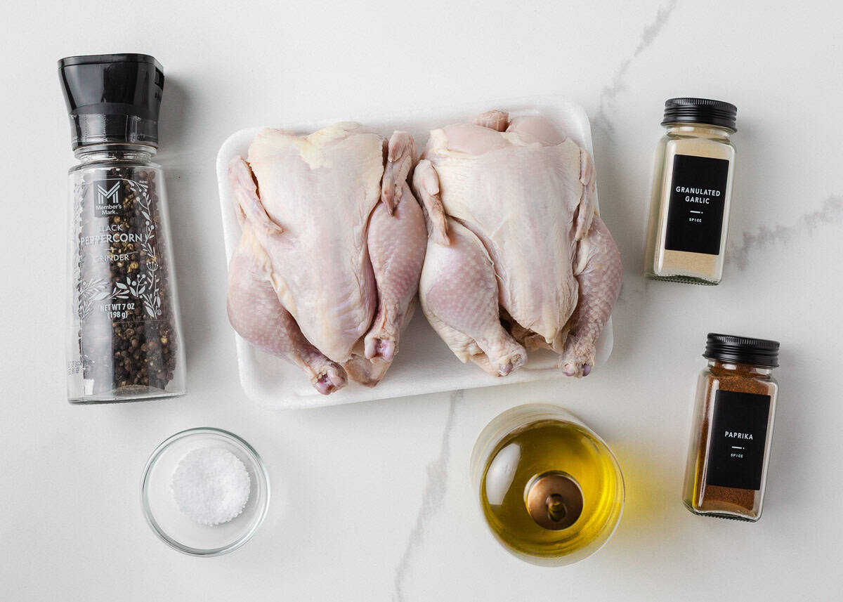 Raw cornish game hens on a tray surrounded by simple ingredients including olive oil, salt, black pepper, garlic powder, and paprika for roasting.