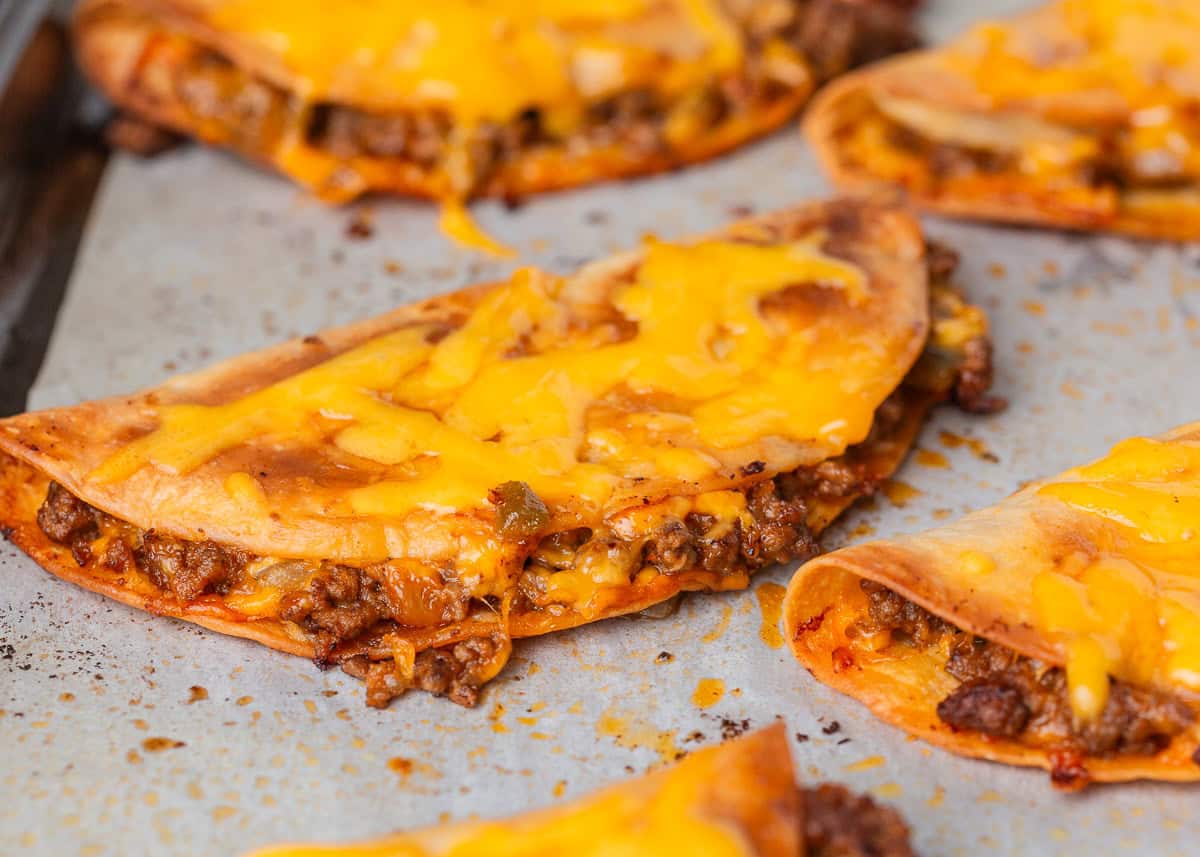 Quick, customizable, and packed with a meaty, cheesy filling, these sheet pan tacos are the perfect family dinner that comes with kid-approved flavor and minimal cleanup! Close-up of crispy sheet pan tacos filled with seasoned ground beef and melted cheddar cheese on a parchment-lined baking sheet.