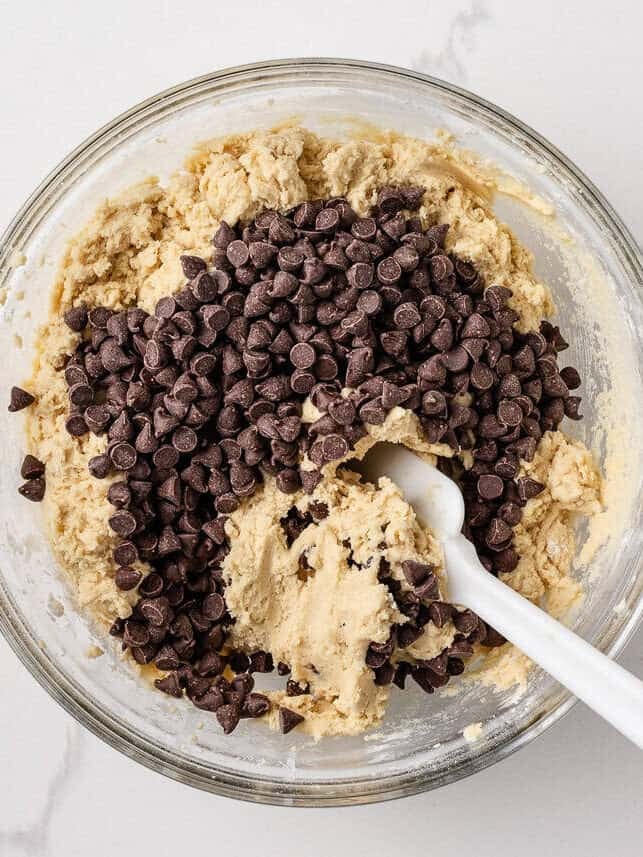 Stirring in chocolate chips into thick cookie dough in a glass bowl, ready to be stirred.