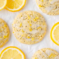 Soft and chewy lemon poppy seed cookies topped with a lemon glaze and zest, surrounded by bright lemon slices on white parchment.