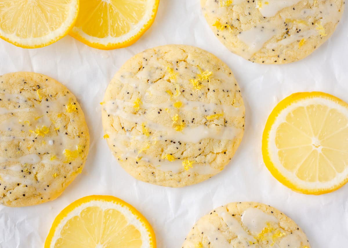 These lemon poppy seed cookies are a no-chill soft, buttery sugar cookie studded with crunchy poppy seeds, infused with sweet citrus flavor, and topped with tangy lemon glaze. Soft and chewy lemon poppy seed cookies topped with a lemon glaze and zest, surrounded by bright lemon slices on white parchment.