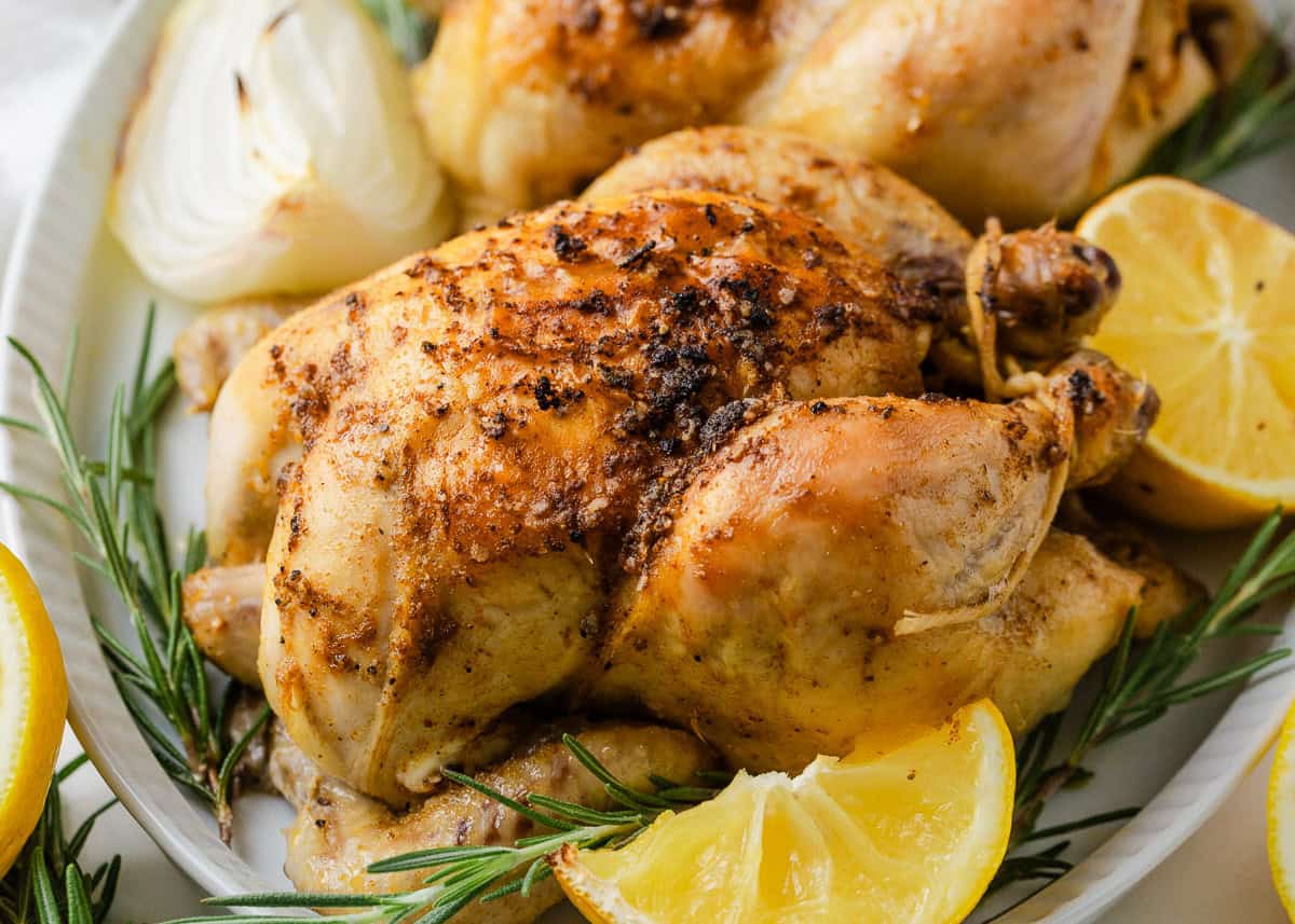 Baked cornish hens with crispy seasoned skin, garnished with lemon wedges, onion and rosemary.