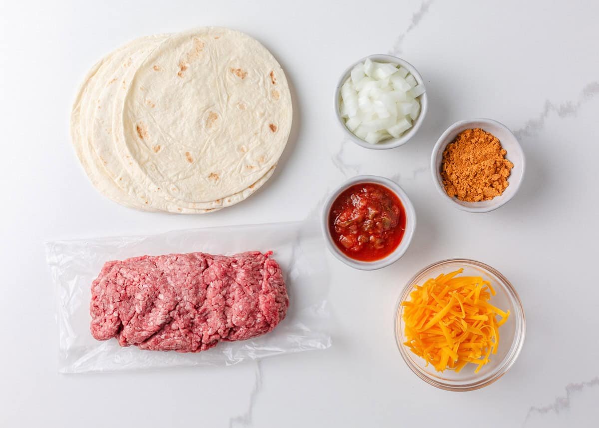 Ingredients for sheet pan tacos including flour tortillas, ground beef, diced onion, taco seasoning, salsa, and shredded cheddar cheese.