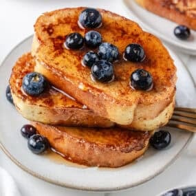 Stack of sourdough French toast slices topped with blueberries and maple syrup on a white plate.