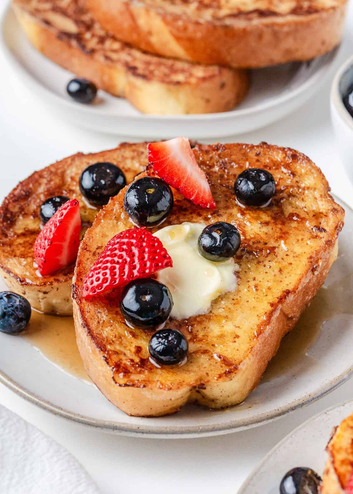Thick slices of sourdough French toast topped with butter, blueberries, strawberries, and syrup on a breakfast plate.