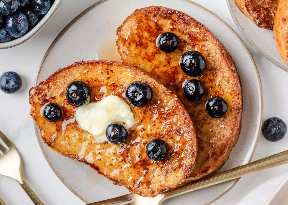 Two slices of delicious sourdough French toast topped with butter, fresh blueberries, and syrup on a white plate.