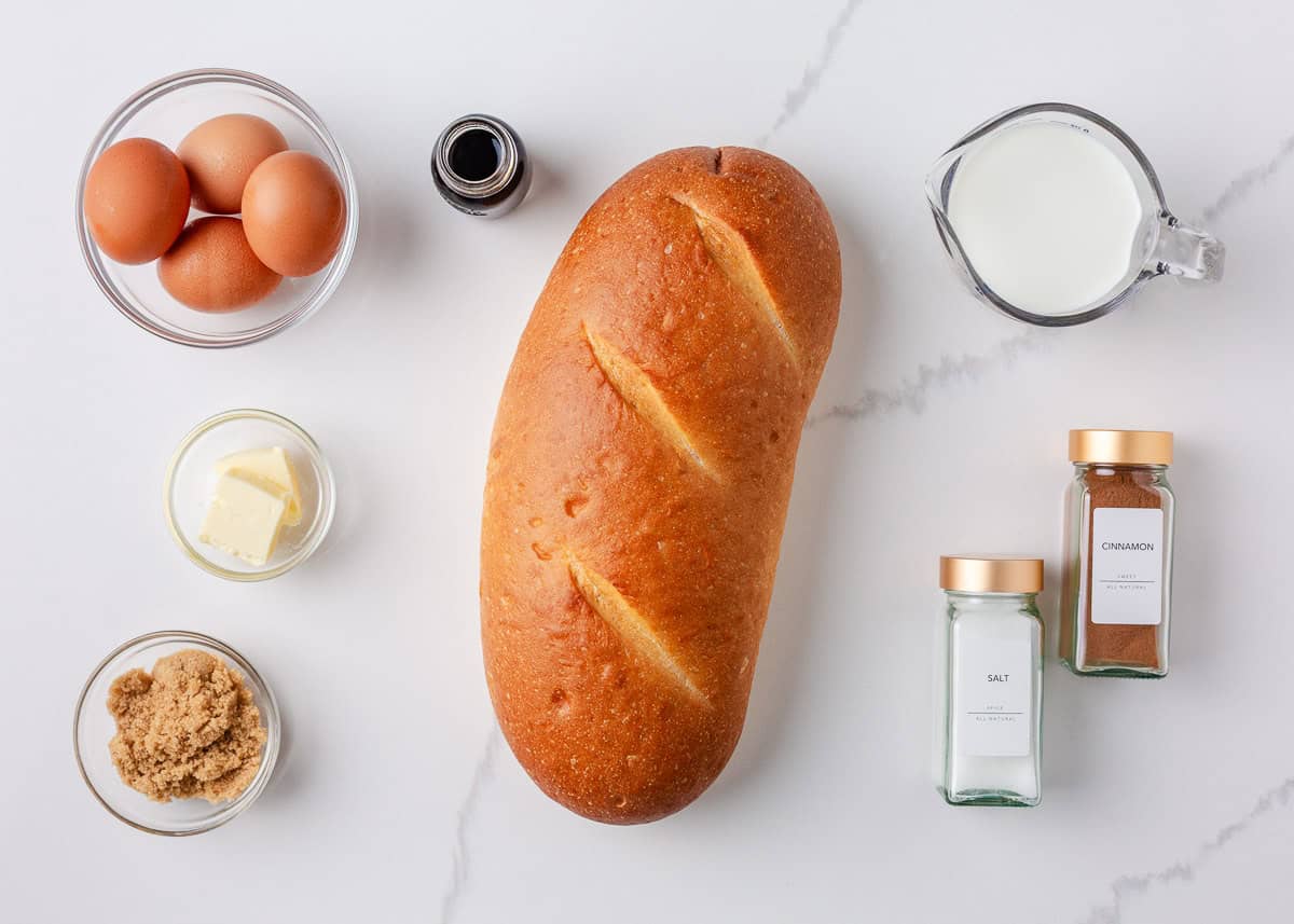 Ingredients for sourdough French toast including fresh bread, eggs, milk, cinnamon, vanilla, butter, and brown sugar on a white surface.