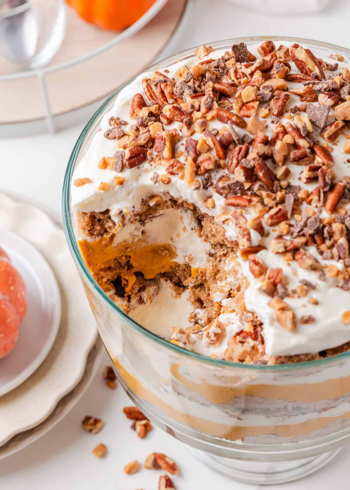 Pumpkin trifle with layers of pumpkin cheesecake filling, whipped cream, spice cake, and pecans in a glass trifle bowl with a scoop missing.