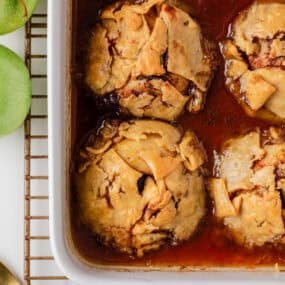 Freshly baked apple dumplings in a white casserole dish, bubbling in a cinnamon brown sugar sauce.