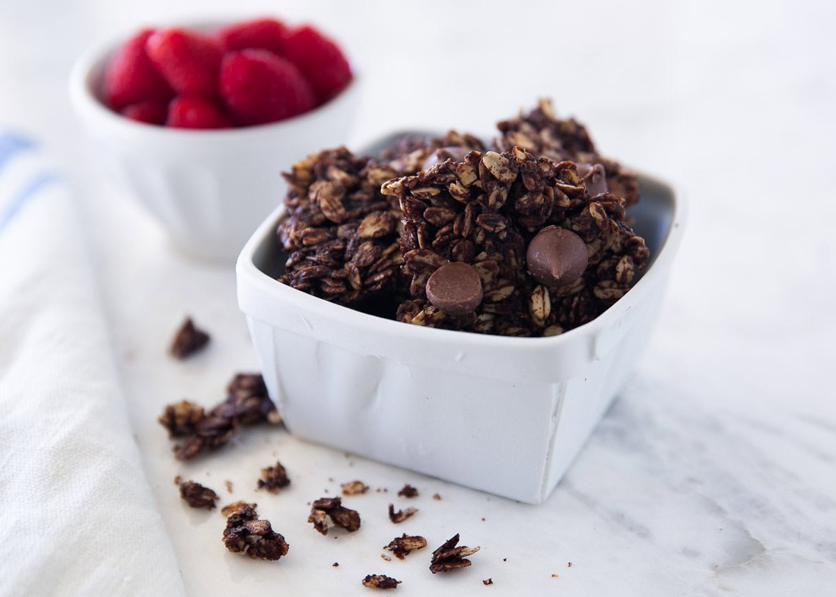Chocolate granola in a bowl square white bowl with raspberries in the background.