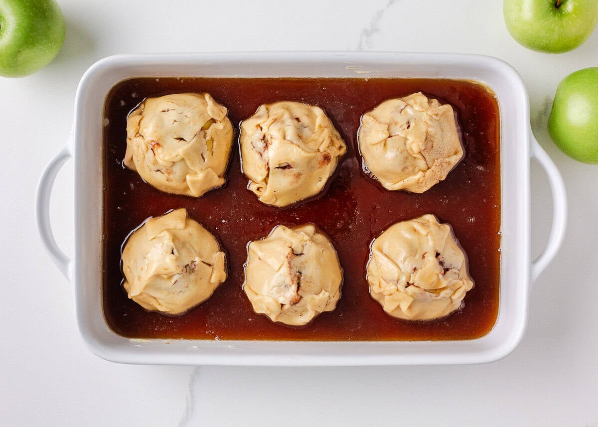 Unbaked apple dumplings wrapped in dough and sitting in brown sugar sauce inside a white baking dish.