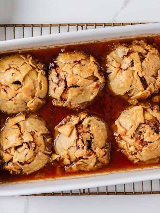 Freshly baked old fashioned apple dumplings golden brown and bubbling in syrup inside a casserole dish.