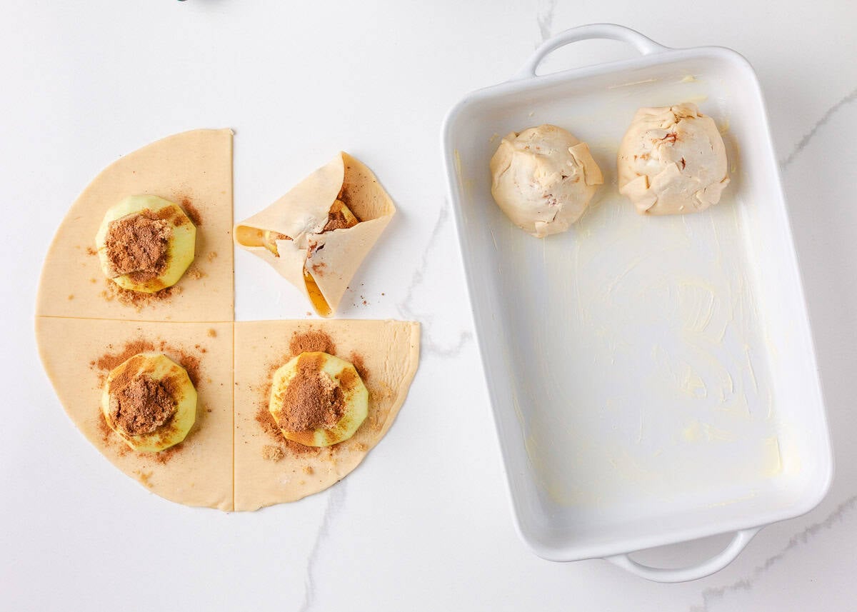 Partially assembled apple dumplings being wrapped in dough and placed into a buttered baking dish.