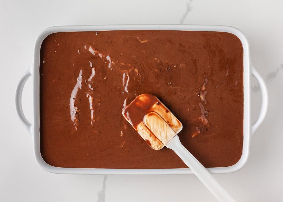 Spreading chocolate ganache over cake in a white baking dish.