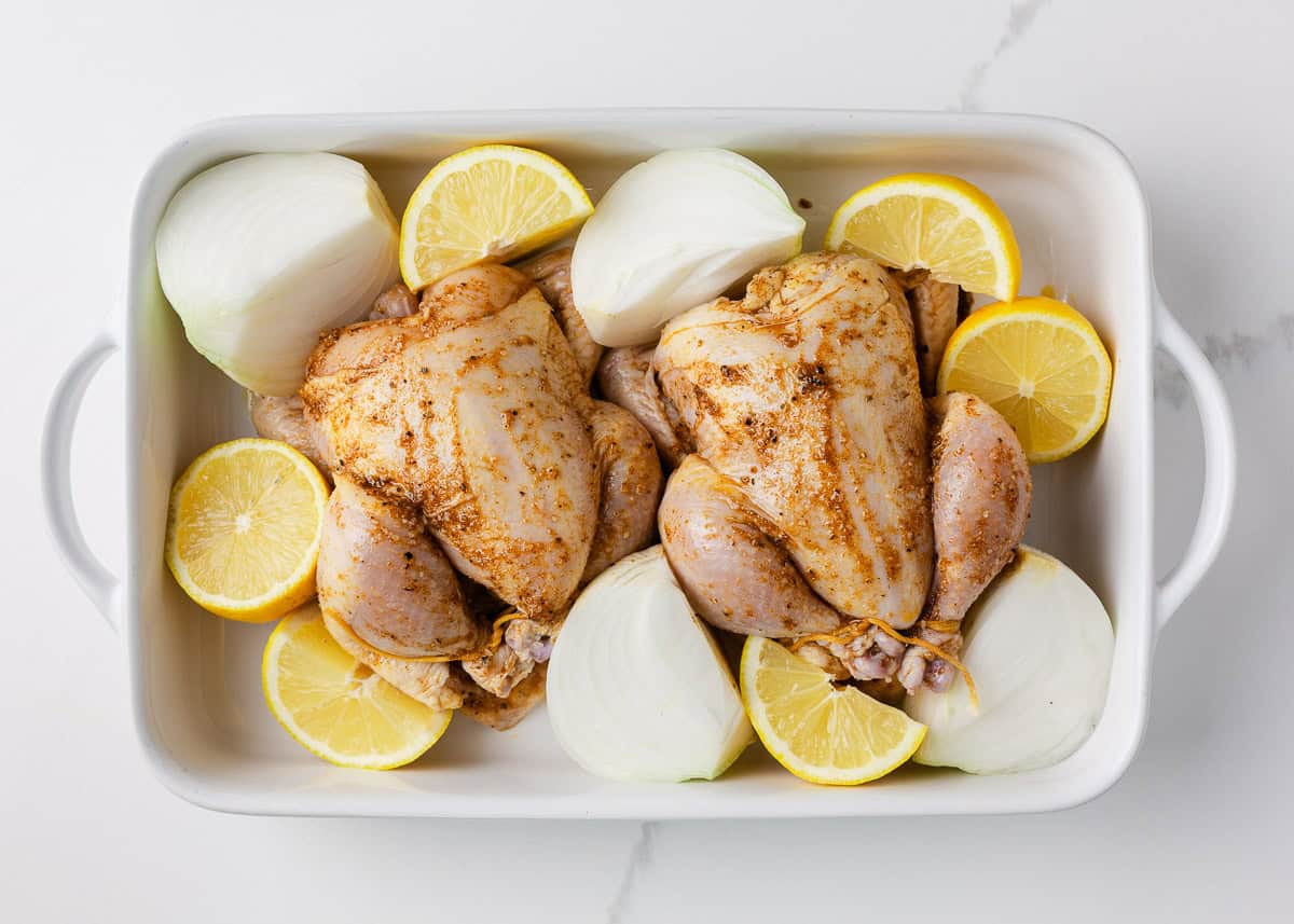 Two seasoned cornish game hens placed in a baking pan with lemon and onion.