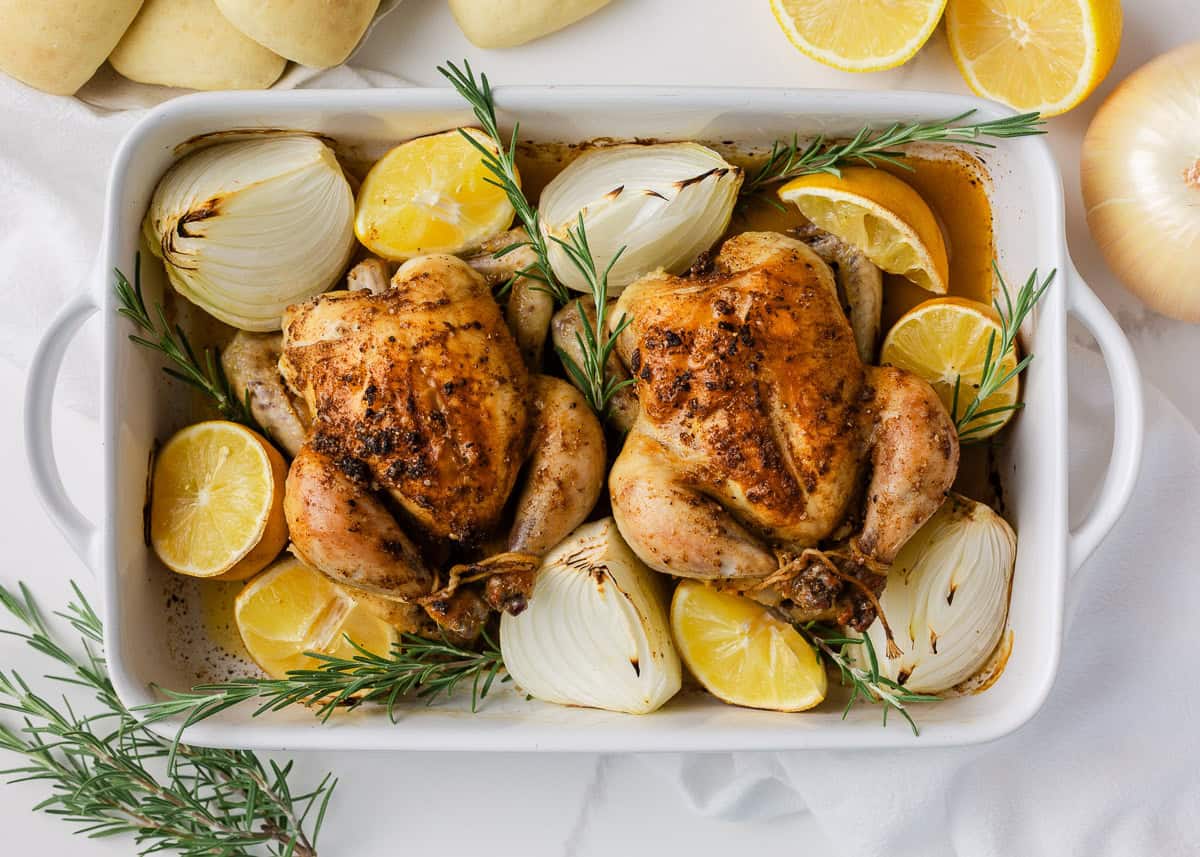 Pan of roasted cornish hens with golden seasoned skin, lemon and onion around the edges, garnished with fresh rosemary.