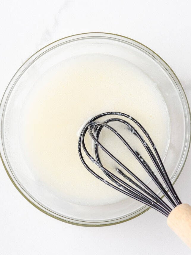 These lemon poppy seed cookies are a no-chill soft, buttery sugar cookie studded with crunchy poppy seeds, infused with sweet citrus flavor, and topped with tangy lemon glaze. Powdered sugar, lemon juice, and milk being whisked into a smooth glaze in a glass bowl.