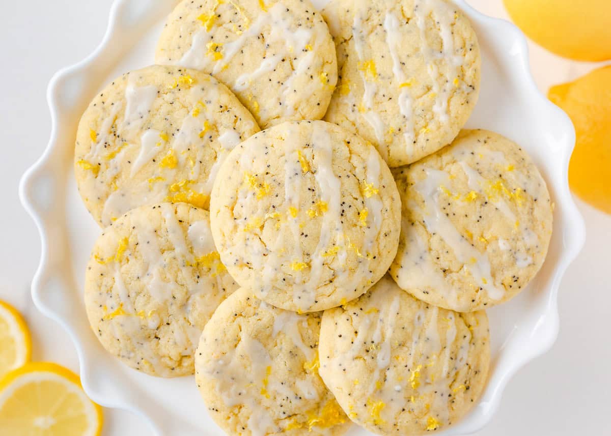 These lemon poppy seed cookies are a no-chill soft, buttery sugar cookie studded with crunchy poppy seeds, infused with sweet citrus flavor, and topped with tangy lemon glaze. A plate stacked with soft lemon poppy seed cookies drizzled with glaze and sprinkled with lemon zest, ready to serve.