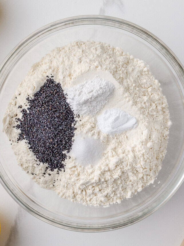 These lemon poppy seed cookies are a no-chill soft, buttery sugar cookie studded with crunchy poppy seeds, infused with sweet citrus flavor, and topped with tangy lemon glaze. Flour, baking soda, baking powder, salt, and poppy seeds added to a mixing bowl, ready to be whisked together.