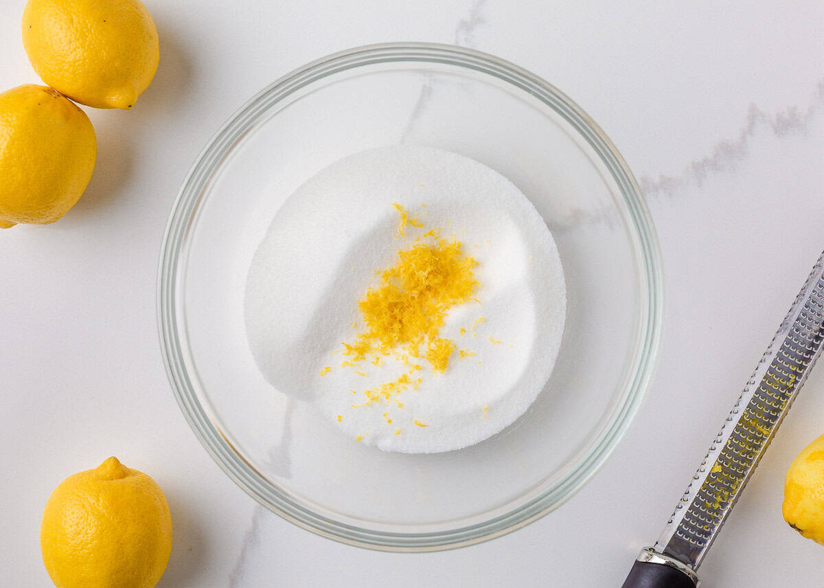 These lemon poppy seed cookies are a no-chill soft, buttery sugar cookie studded with crunchy poppy seeds, infused with sweet citrus flavor, and topped with tangy lemon glaze. Granulated sugar and freshly grated lemon zest in a glass bowl, ready to be rubbed together for fragrant lemon sugar.