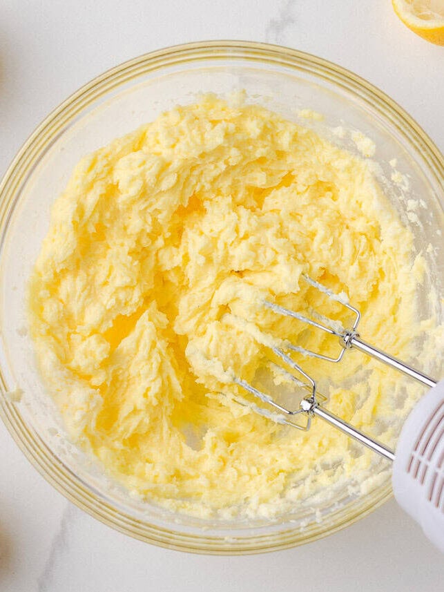 These lemon poppy seed cookies are a no-chill soft, buttery sugar cookie studded with crunchy poppy seeds, infused with sweet citrus flavor, and topped with tangy lemon glaze. Butter, sugar, lemon zest, and lemon juice creamed together in a glass bowl with a hand mixer, forming a fluffy lemon base.