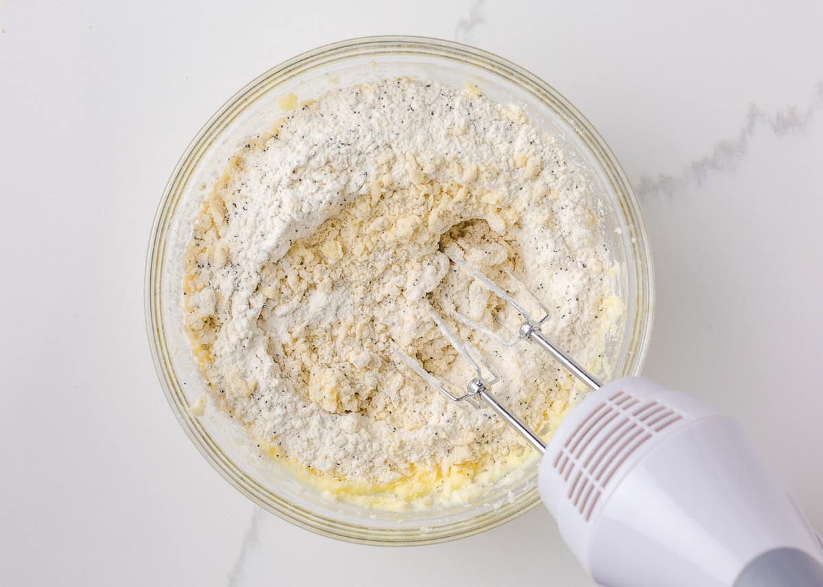 These lemon poppy seed cookies are a no-chill soft, buttery sugar cookie studded with crunchy poppy seeds, infused with sweet citrus flavor, and topped with tangy lemon glaze. Dry ingredients partially mixed into the creamed lemon butter mixture using a hand mixer in a large bowl.