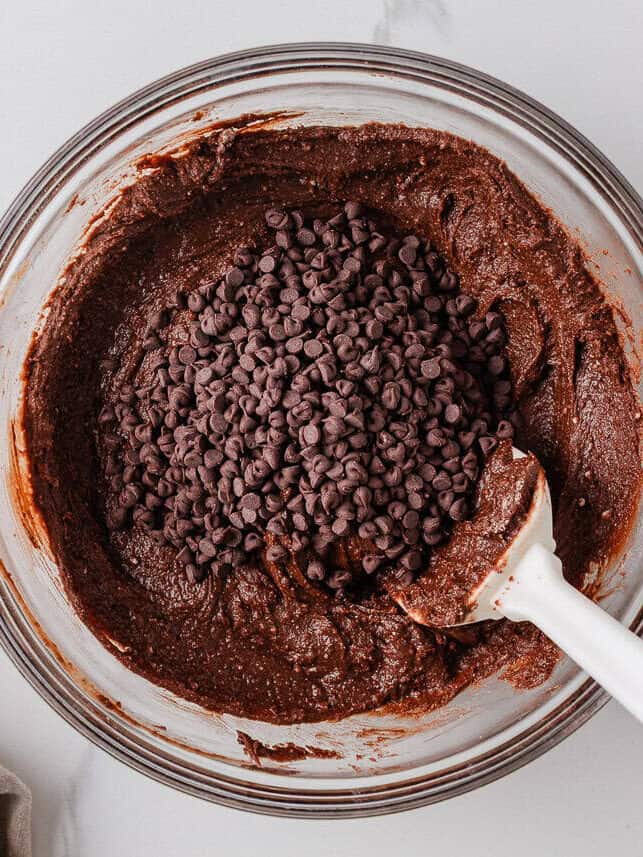 Chocolate brownie batter in a mixing bowl topped with mini chocolate chips and a spatula for stirring.