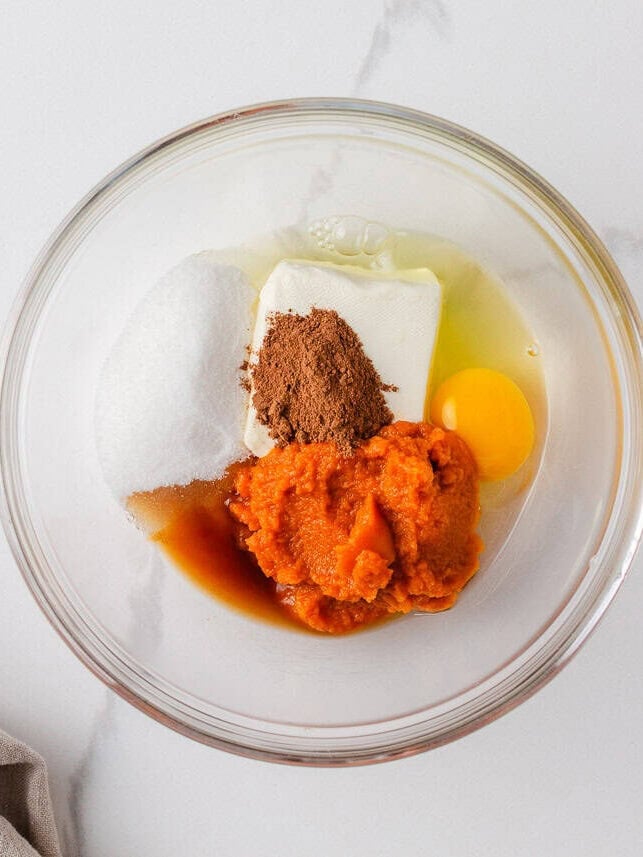 Pumpkin brownie cheesecake layer ingredients in a mixing bowl, including pumpkin puree, cream cheese, sugar, egg, cinnamon, and vanilla.