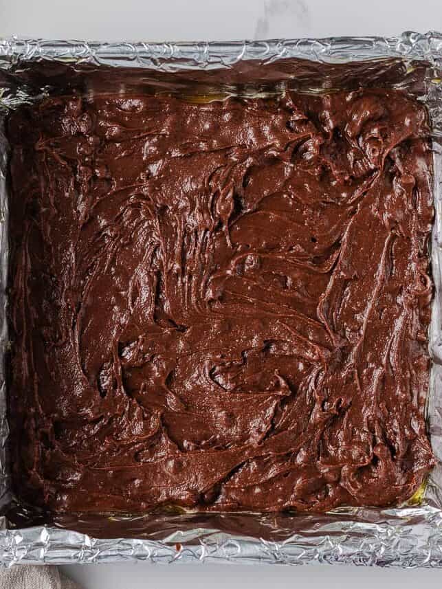 Foil-lined baking pan filled with chocolate brownie batter, ready for the pumpkin layer.