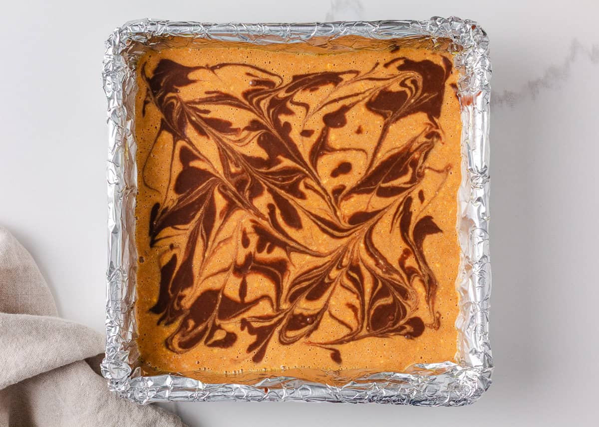 Unbaked pumpkin brownies in a foil-lined pan with a marbled chocolate and pumpkin swirl pattern.